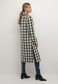 Abrigo a cuadros en blanco y negro con corte recto, aberturas laterales, detalles de botones, combinado con jeans azules y botines negros.