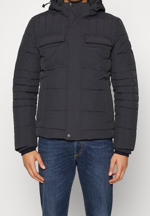 Homme portant une veste d'hiver noire matelassée avec poches poitrine à rabat, fermeture éclair, et un jean bleu, debout devant un fond blanc.