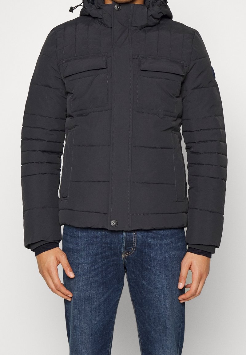 Homme portant une veste d'hiver noire matelassée avec poches poitrine à rabat, fermeture éclair, et un jean bleu, debout devant un fond blanc.