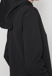 Sudadera con capucha negra de tela suave y texturizada. Presenta una capucha grande y mangas ajustadas, mostrando una vista lateral de la prenda.