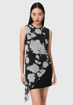 Młoda kobieta ubrana w czarną, bezrękawnikową sukienkę z dużym białym kwiatowym wzorem i asymetrycznym dołem, stojąca na jednolitym jasnym tle.