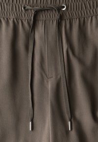Brown drawstring pants feature an elastic waistband with silver hardware, smooth fabric texture, and a single stitched pocket detail.
