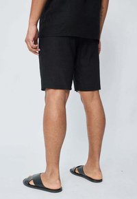 Shorts en lin noirs à coupe décontractée, arrivant à mi-cuisse, accompagnés de sandales à glissière noires. Design et texture simples, sans ornement.