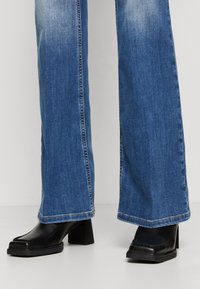 Svarta klackade stövlar med fyrkantig tå, tillsammans med vida blå denimjeans med en blekt tvätt och fransad fåll.