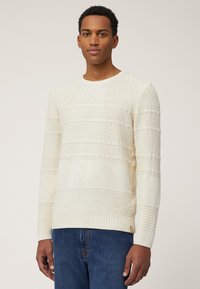 Cremefarbener Pullover mit strukturierten Streifen, der verschiedene gestrickte Muster aufweist. Runder Ausschnitt und gerippte Bündchen, kombiniert mit blauen Jeans.