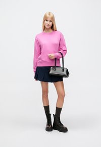 Ροζ φούτερ, ναυτικό μπλε φούστα, μαύρη τσάντα χειρός με λαβές, καφέ κάλτσες μέχρι το γόνατο και μαύρα μποτάκια με κορδόνια και μαύρη χοντρή σόλα. Απλό φόντο.