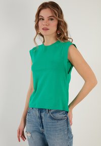 Top verde senza maniche realizzato in morbido tessuto, con scollatura rotonda e vestibilità comoda, abbinato a jeans azzurri chiari strappati.