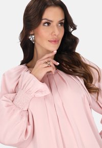 Rosa Bluse mit Falten, Puffärmeln und gesmokten Bündchen. Das Model trägt auffällige silberne Statement-Ohrringe und einen goldenen geometrischen Ring.