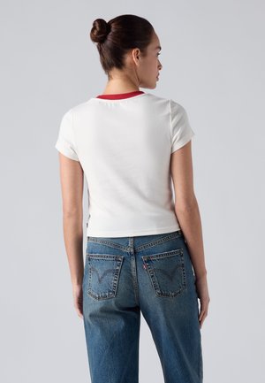 Femme aux cheveux attachés en chignon portant un t-shirt blanc à manches courtes avec un col rouge et un jean bleu, de dos sur un fond gris.