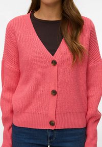 Žena nosi debeli ružičasti pleteni kardigan s tri velika smeđa gumba preko crne majice i plavih traperica, vidljiv struk i torzo.