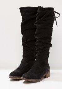 Botas negras de ante por encima de la rodilla con un diseño holgado, tacón plano y suela texturizada. Cuentan con una cremallera lateral y detalles de lazo en la parte superior.