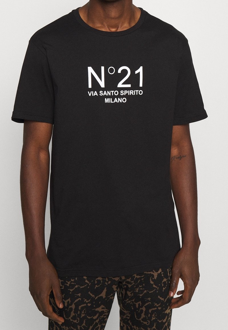 Czarny t-shirt z białym napisem "N°21 VIA SANTO SPIRITO MILANO" noszony przez osobę w brązowych spodni z wzorem i tatuażem na lewym przedramieniu.