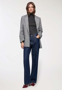 Donna che indossa un blazer grigio a quadri, un dolcevita nero, jeans a gamba larga blu scuro e tacchi a punta bordeaux, in piedi con una mano in tasca.