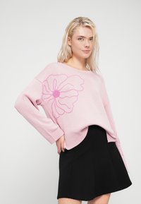 Ροζ πλεκτό πουλόβερ με φαρδιά εφαρμογή, ριμπ μανσέτες και πλευρικά σκισίματα. Φέρει μεγάλο ροζ λουλούδι στο μπροστινό μέρος. Συνδυασμένο με μαύρη φούστα.