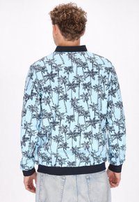 Giacca bomber di colore blu cielo con stampa nera di palme, con polsini e orlo a costine e colletto nero. Tessuto liscio con materiale leggero.