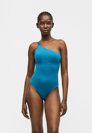 Femeie purtând un costum de baie teal cu un umăr gol, stând în picioare pe un fundal simplu deschis, privind înainte cu o expresie neutră.