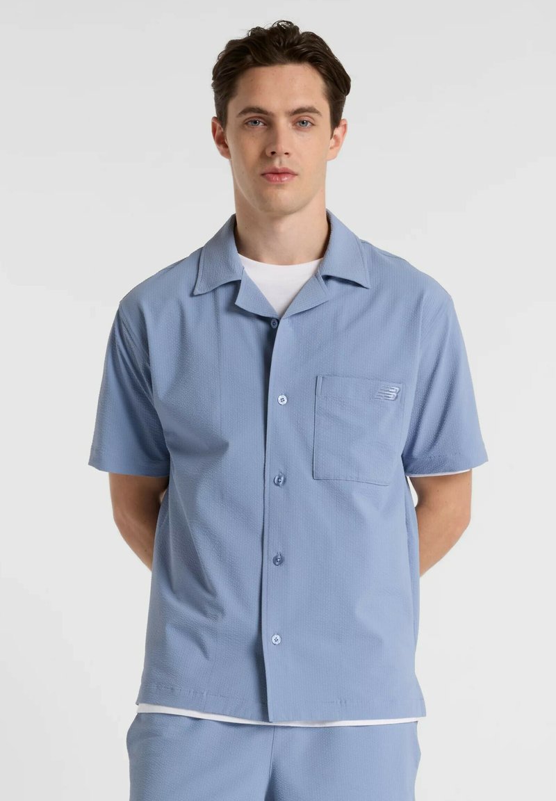 Chemise bleu clair à manches courtes avec un tissu texturé, fermeture boutonnée sur le devant, une poche poitrine unique et un logo subtil sur la poche.