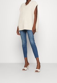 Krämfärgad stickad oversized väst med V-ringning, tillsammans med blå denim skinny jeans med fransat avslut, och vita öppna högklackade mules.