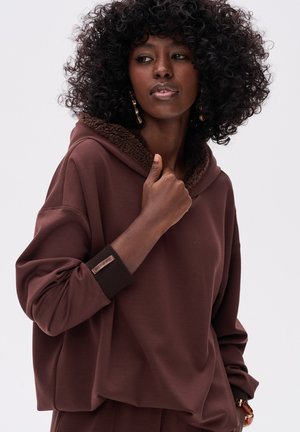 Brązowa, oversize'owa bluza z kapturem z pluszowym, brązowym wykończeniem na podszewce. Posiada luźne rękawy oraz teksturowany mankiet z logo.