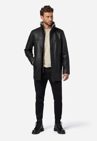 Schwarze Lederjacke mit hohem Kragen, Knopfverschluss und zwei seitlichen Taschen, getragen über einem hellen Pullover und kombiniert mit schwarzen Hosen und Stiefeln.