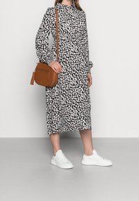 Robe midi noire à imprimé floral avec des manches longues et une taille froncée, accompagnée d'un sac bandoulière beige et de baskets blanches sur un fond gris.