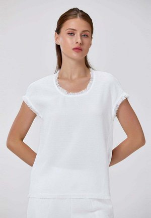 Jeune femme aux cheveux châtain clair portant un haut blanc à manches courtes texturé avec des bords effilochés, posant devant un fond clair uni.