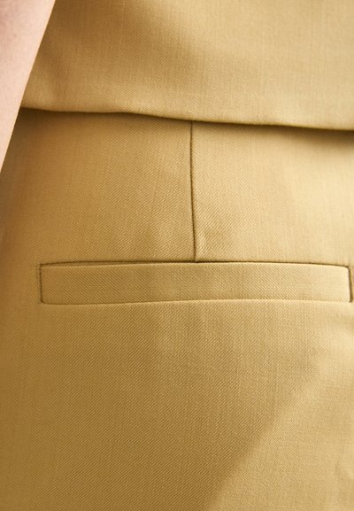 Tessuto giallo chiaro con una texture liscia, caratterizzato da una piccola tasca piatta e dettagli delle cuciture puliti. Il design è minimalista e sartoriale.