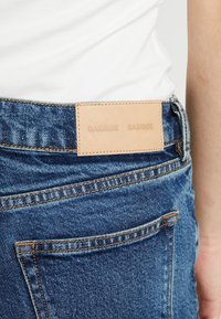 Vaqueros de mezclilla azul con un parche de cuero marrón claro en la cintura. Detalles de costura en contraste y un bolsillo trasero visible.