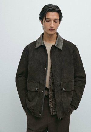 Giacca in suede marrone con colletto grigio, tasche frontali e bottoni a pressione. Texture liscia e vestibilità rilassata. Indossata sopra un maglione lavorato a maglia.