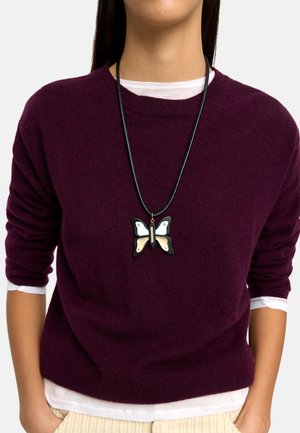 Czarny skórzany naszyjnik z wisiorkiem w kształcie motyla z białymi, złotymi i czarnymi akcentami. Modelka ubrana w fioletowy sweter na białej koszuli.