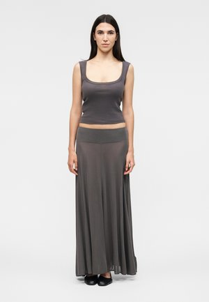 Νέα γυναίκα με μακριά σκούρα μαλλιά στέκεται μπροστά φορώντας ένα γκρι αμάνικο πάνω μέρος και μια αέρινη μακριά γκρι φούστα με μαύρα παπούτσια.