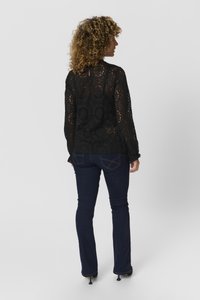 Blusa nera in pizzo con intricati motivi floreali, maniche lunghe con polsini a ruches e collo alto. Abbinata a jeans svasati blu scuro.