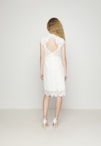Robes en dentelle blanche à manches courtes, dos nu et ourlet festonné. Présente des motifs floraux complexes et un design jusqu'aux genoux. Portée avec des talons blancs.