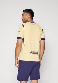 Beige sportsjersey med tekstureret stof, sorte skulderaccenter og blå kant. Har UNHCR-logoet og et lille flag på bagsiden.