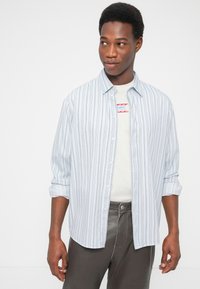 Νεαρός άνδρας που φοράει ένα ανοιχτό μπλε ριγέ πουκάμισο πάνω από λευκό μπλουζάκι με σχέδιο και σκούρο παντελόνι, κοιτώντας προς τα αριστερά του.