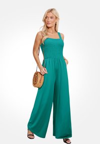 Teal jumpsuit met een gerimpelde lijf, wijde benen en verstelbare bandjes; gecombineerd met een ronde gevlochten handtas en zwarte schuifsandalen.