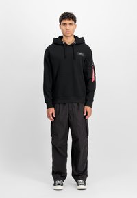 Sudadera con capucha negra, con un bolsillo frontal y un parche de logo, combinada con pantalones cargo negros y zapatillas negras con suelas blancas.