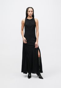 Vestido maxi preto sem mangas com superfície texturizada, apresentando uma fenda lateral alta e um decote franzido. Combinado com botas pretas de salto fino pontiagudas.