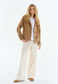 Žena s dlhými blond vlasmi, ktorá má na sebe hnedú zapínaciu bundu, bielu košeľu, krémové široké nohavice a hnedé topánky, stojaca na bielom pozadí.