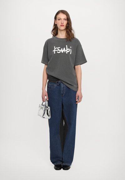 Camiseta gris de gran tamaño con logo blanco, combinada con jeans de mezclilla azul de pierna ancha. Los accesorios adicionales incluyen un bolso transparente y zapatos negros.