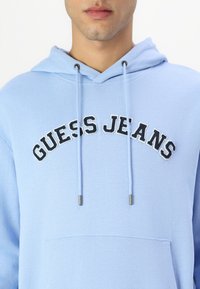 Sudadera con capucha de color azul claro hecha de un tejido suave, con el logo "GUESS JEANS" bordado en negro sobre un bolsillo tipo canguro. Cuerdas con punta de plata.