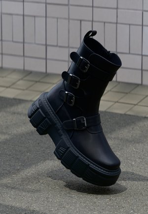 Bottine noire plateforme épaisse avec quatre brides à boucle et une semelle épaisse texturée sur un trottoir extérieur près d’un mur carrelé.