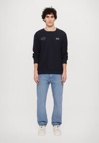 Jūras spēku svītrainā krekla ar apaļu kaklu, garām piedurknēm un zeltīti izšūtu EA7 logotipu uz krūtīm, saskaņots ar gaiši ziliem džinsiem un bēšiem sporta apaviem.