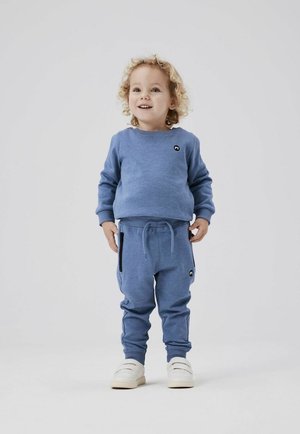 Sudadera azul y joggers a juego, hechos de una tela suave. La sudadera tiene un pequeño logo negro, mientras que los joggers cuentan con bolsillos laterales. Zapatillas blancas.