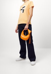 Pomarańczowa skórzana torebka w kształcie półksiężyca z frędzelkami, zestawiona z jasnopomarańczowym t-shirtem z grafiką i ciemnoniebieskimi spodniami typu wide-leg.