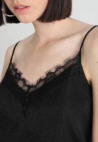 Top nero in satin con spalline regolabili, caratterizzato da un bordo in pizzo e bottoni decorativi sul collo. Texture liscia.