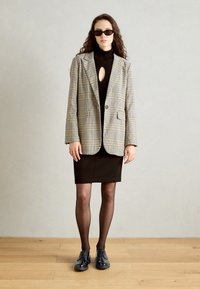 Blazer a scacchi beige e nero sopra un dolce di collo alto marrone con dettaglio a oblò, abbinato a una gonna marrone attillata e scarpe nere con lacci.