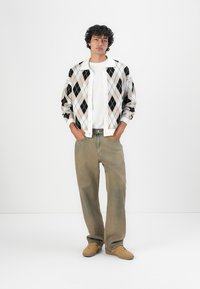 White and black argyle cardigan over a white shirt, paired with loose beige pants and tan sneakers. Model stands with hands in pockets.