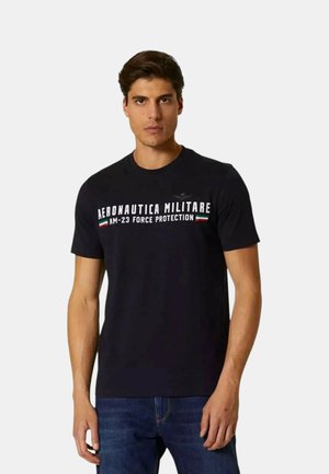 Giovane uomo che indossa una maglietta nera con la scritta "Aeronautica Militare AM-23 Force Protection" e piccole strisce con la bandiera italiana, abbinata a jeans blu.