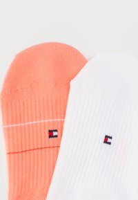 Ζευγάρι κάλτσες: η μία κοραλλί με ραβδωτή υφή και κόκκινο/μπλε λογότυπο, η άλλη λευκή με κάθετες ρίγες και λογότυπο, από βαμβακερό ύφασμα.